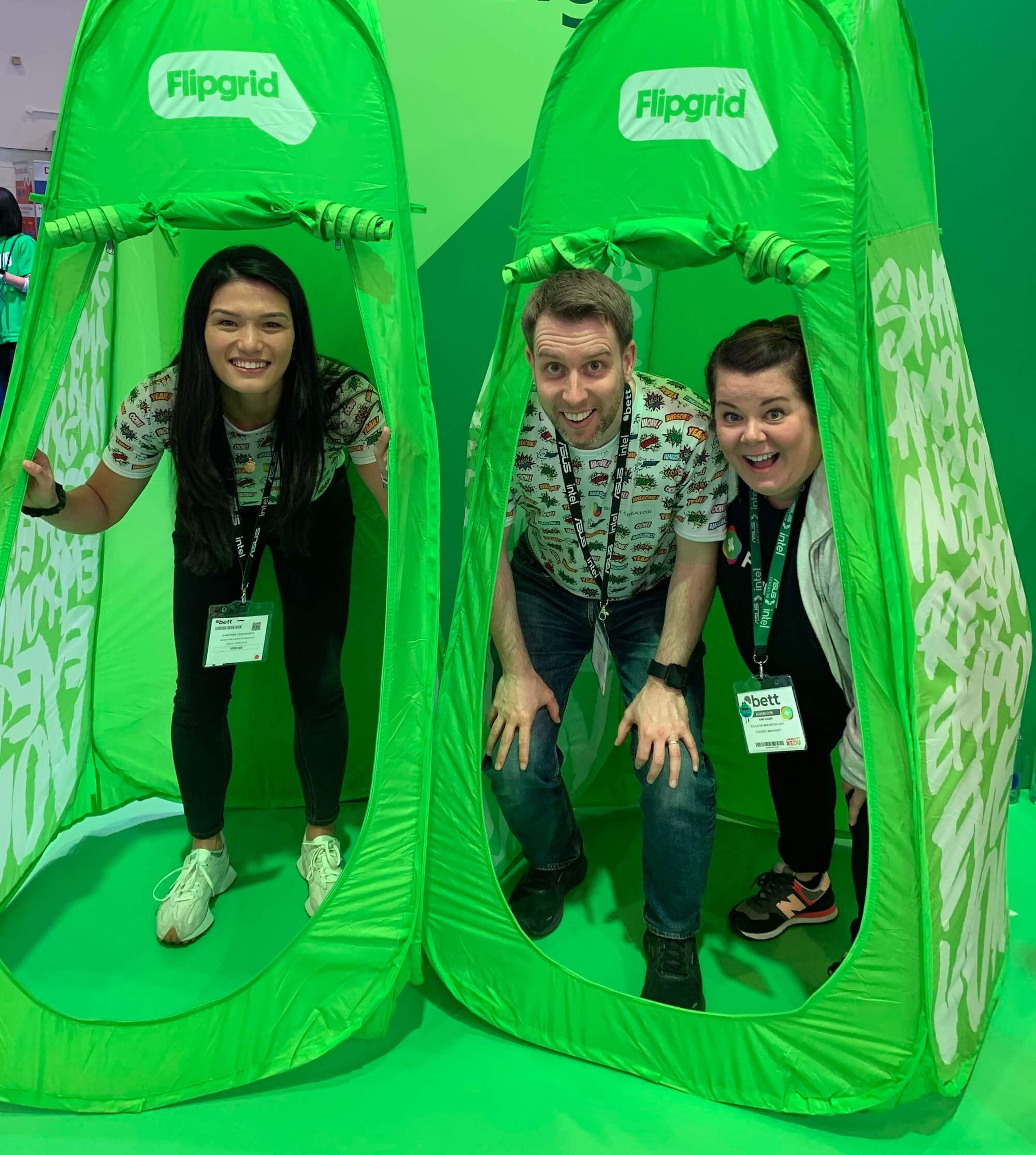 Three people are posing inside bright green pop-up tents with the Flipgrid logo at the top. From left to right, there is a woman with long dark hair, a man with short hair, and a woman with shoulder-length hair. They are all smiling and leaning forward, looking through the openings of the tents. They are wearing badges around their necks, indicating they are at a conference or event. The background is filled with a green color that matches the tents, creating a cohesive and lively atmosphere.