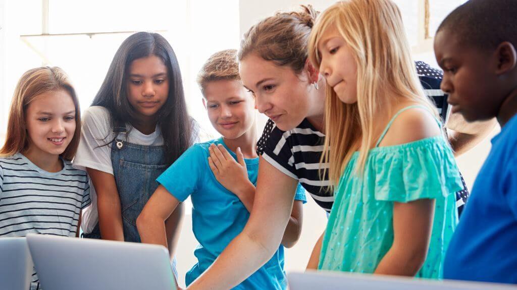 A diverse group of elementary students gathers around a table with laptops, guided by a teacher. The students are engaged and focused, collaborating on a digital project—illustrating inclusive, technology-supported learning in the classroom.