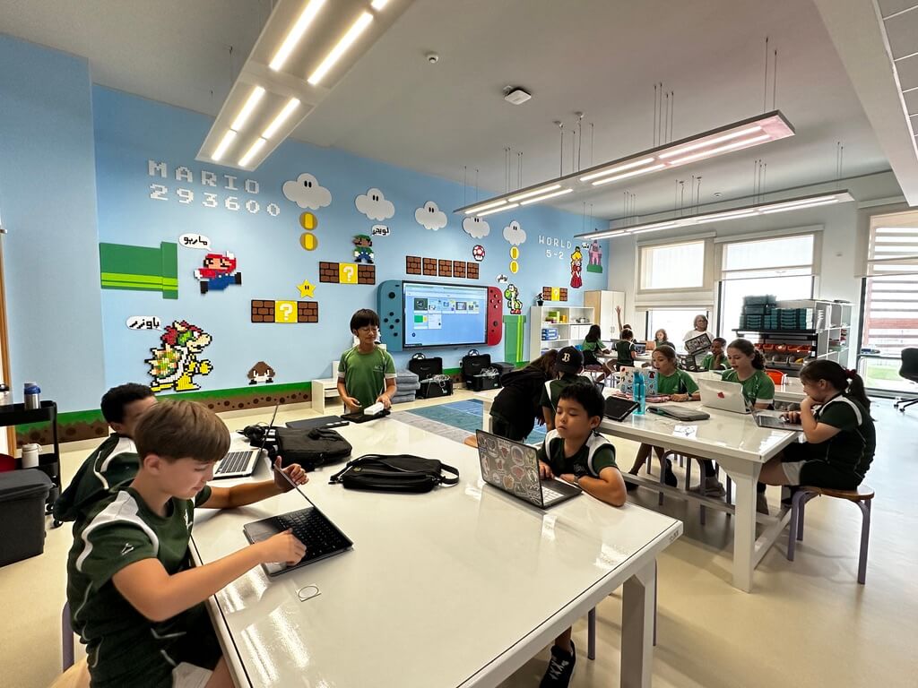 A bright and engaging elementary design classroom filled with students working on laptops at white tables. The students, wearing green school uniforms, are focused and engaged in their tasks. The classroom has a playful Super Mario-themed wall mural, featuring pixelated characters like Mario, Bowser, Goombas, coins, and question blocks. A large screen is mounted on the wall, and various classroom supplies and storage bins are neatly arranged. Natural light pours in from large windows, creating an inviting learning environment.